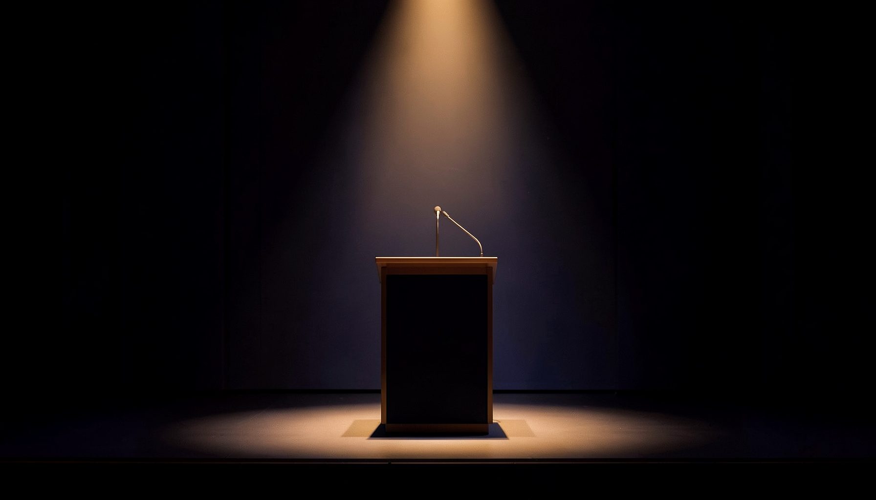 A softly lit conference stage with a single spotlight on an empty lectern, deep navy and warm amber tones, elegant and understated