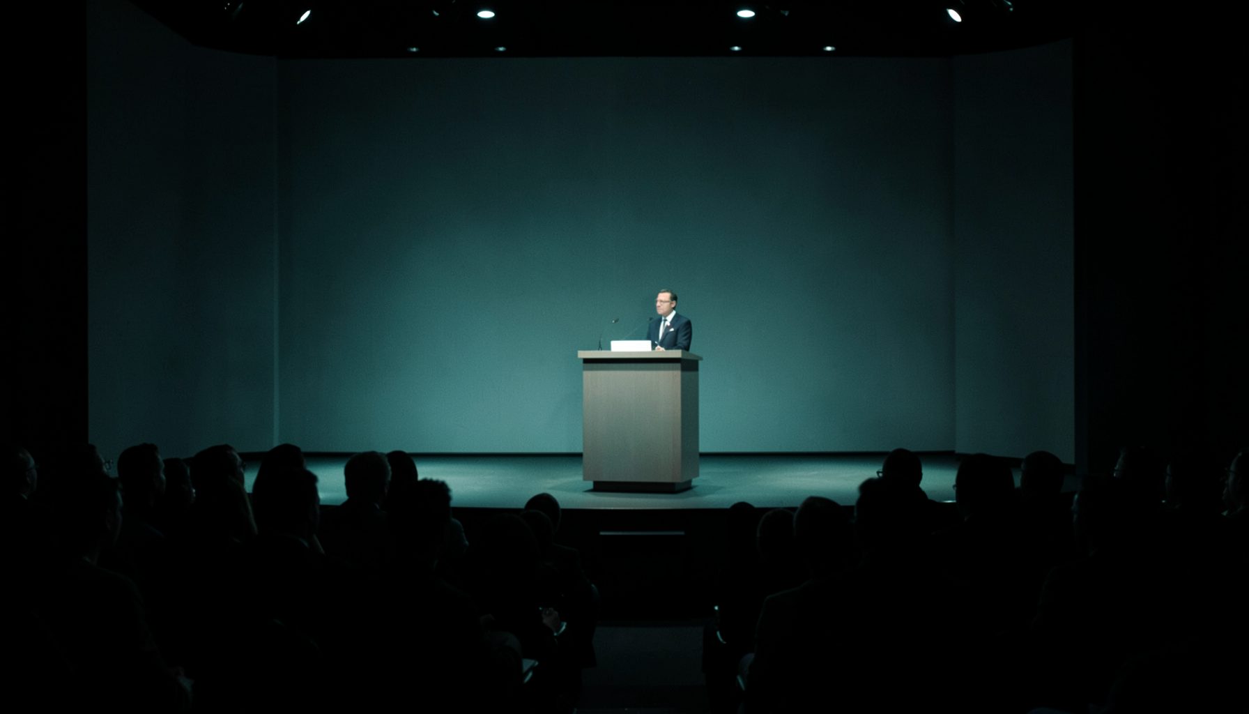 Dimly lit conference stage in muted teal and charcoal shades with a lectern and audience silhouettes, calm and professional mood